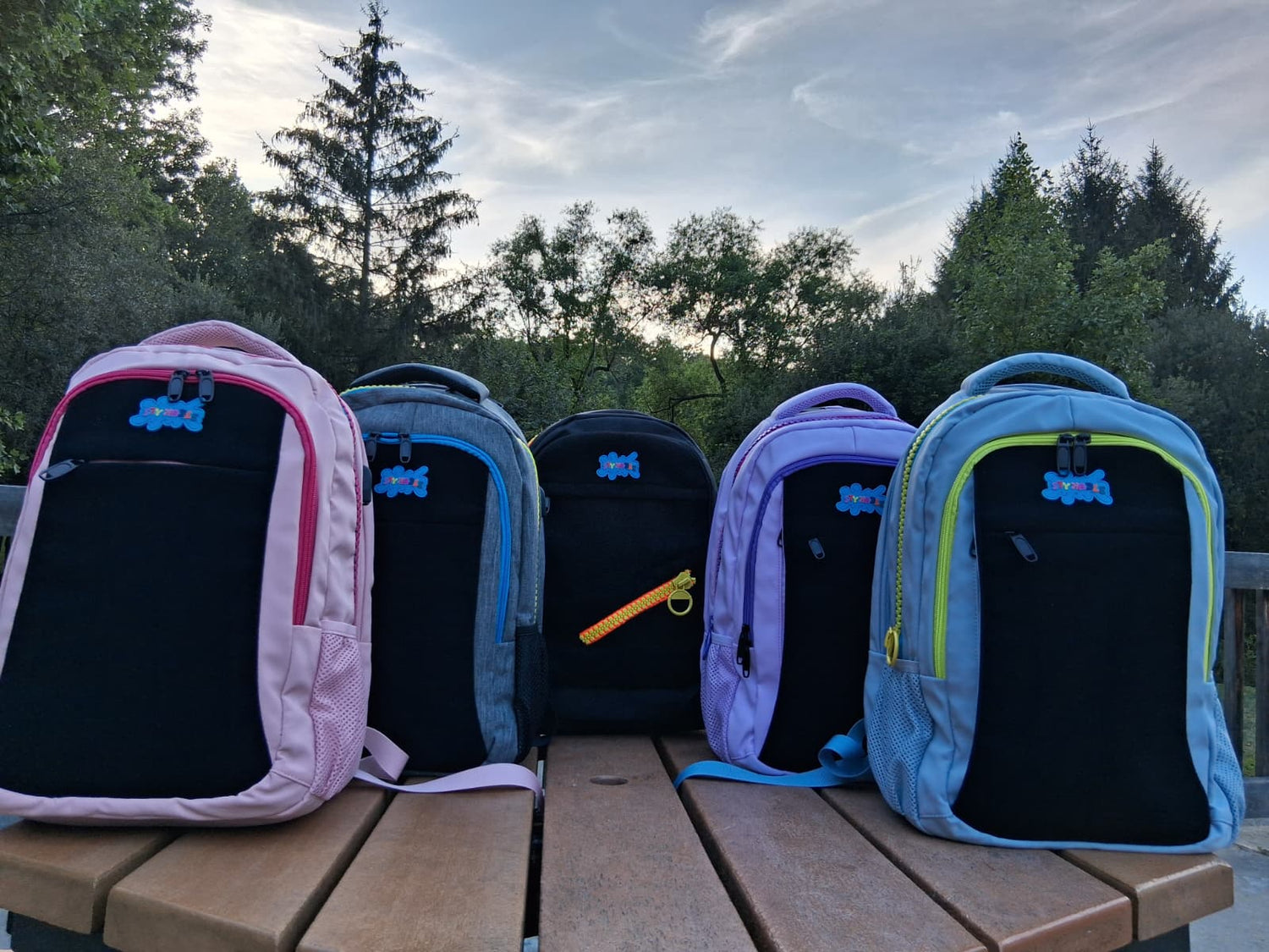 Five colorful backpacks on a wooden surface with trees in the background